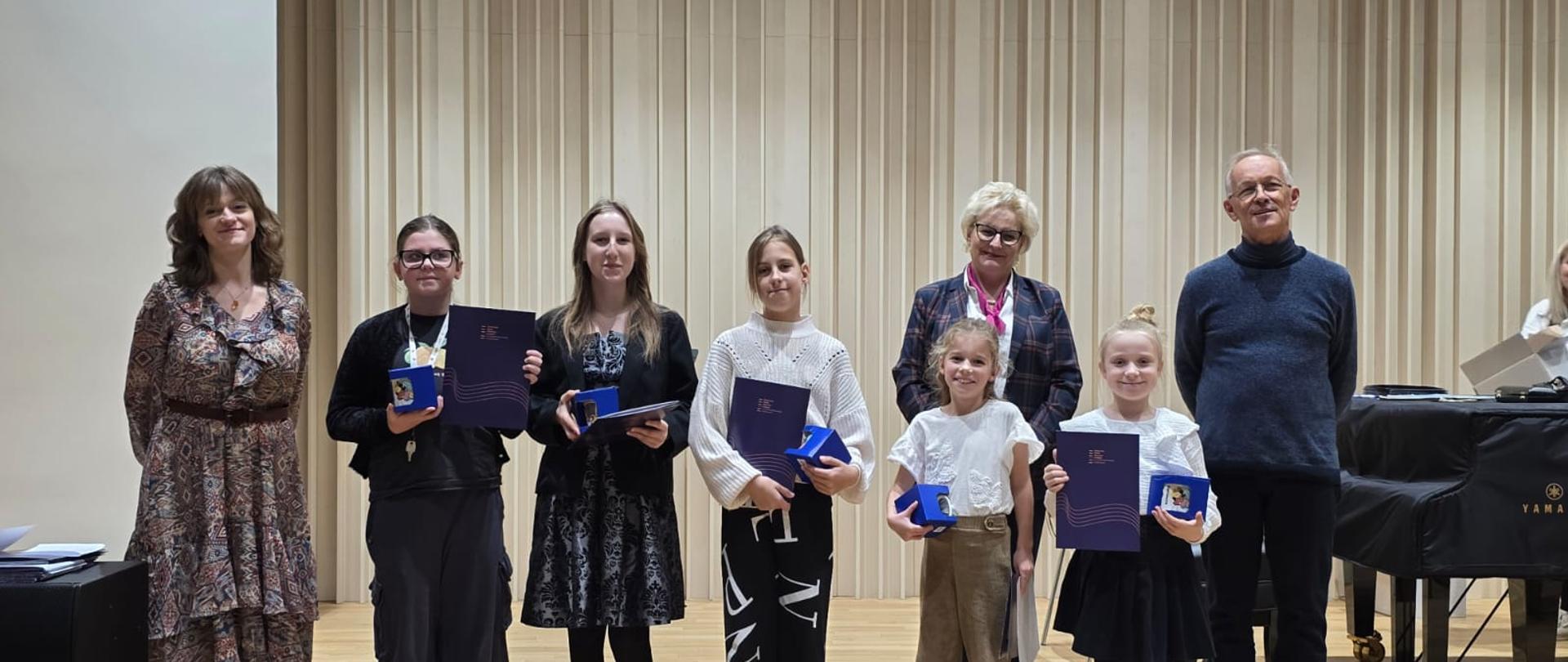 Na zdjęciu grupa uczestników warsztatów muzycznych stoi na scenie w sali koncertowej. W centrum znajdują się uczniowie trzymający dyplomy i upominki, a po bokach stoją pedagodzy prowadzący warsztaty oraz nauczyciele. W tle widoczna jest drewniana akustyczna ściana i fortepian. Wszyscy uśmiechnięci, pozują do pamiątkowego zdjęcia po zakończeniu wydarzenia.