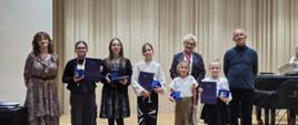 Na zdjęciu grupa uczestników warsztatów muzycznych stoi na scenie w sali koncertowej. W centrum znajdują się uczniowie trzymający dyplomy i upominki, a po bokach stoją pedagodzy prowadzący warsztaty oraz nauczyciele. W tle widoczna jest drewniana akustyczna ściana i fortepian. Wszyscy uśmiechnięci, pozują do pamiątkowego zdjęcia po zakończeniu wydarzenia.