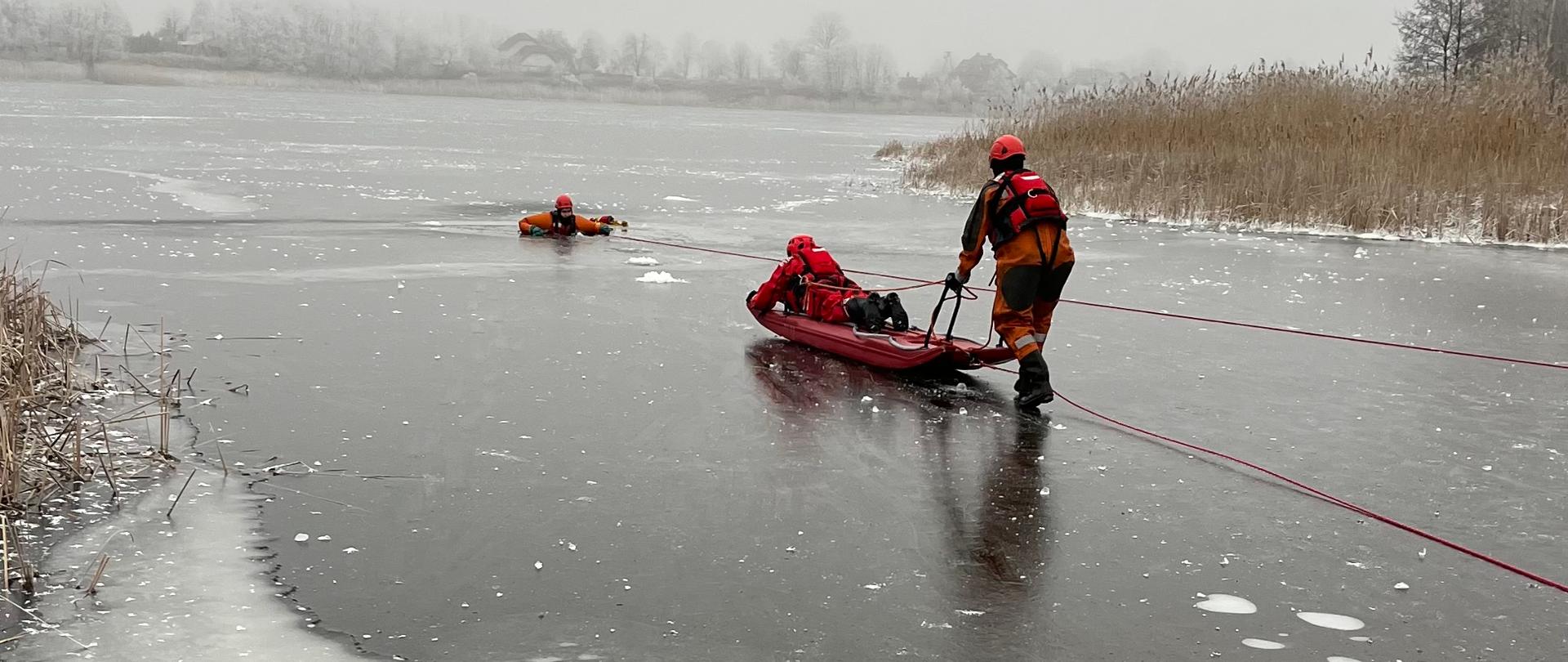 Zdjęcie przedstawia strażaków ćwiczących na lodzie. Strażacy zabezpieczeni w ubrania do pracy w wodzie. Jeden ze strażaków topi się w przeręblu. Dwóch strażaków zabezpieczonych liną, w tym jeden leży na desce lodowej podchodzą do poszkodowanego.