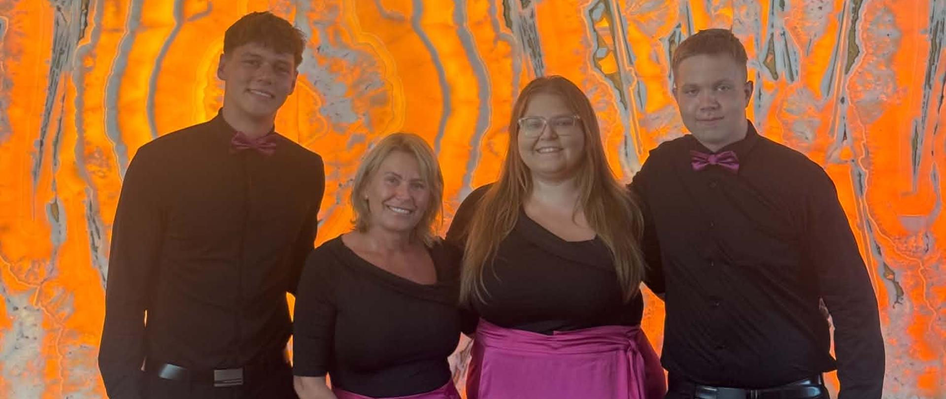 Czworo członków chóru Cantabile Leo Cantores — uczeń, dwoje absolwentów oraz nauczycielka — stoi przed pomarańczowo-szarą, podświetlaną ścianą. Kobiety mają czarne bluzki i różowe spódnice, a mężczyźni czarne stroje z różowymi muszkami.
