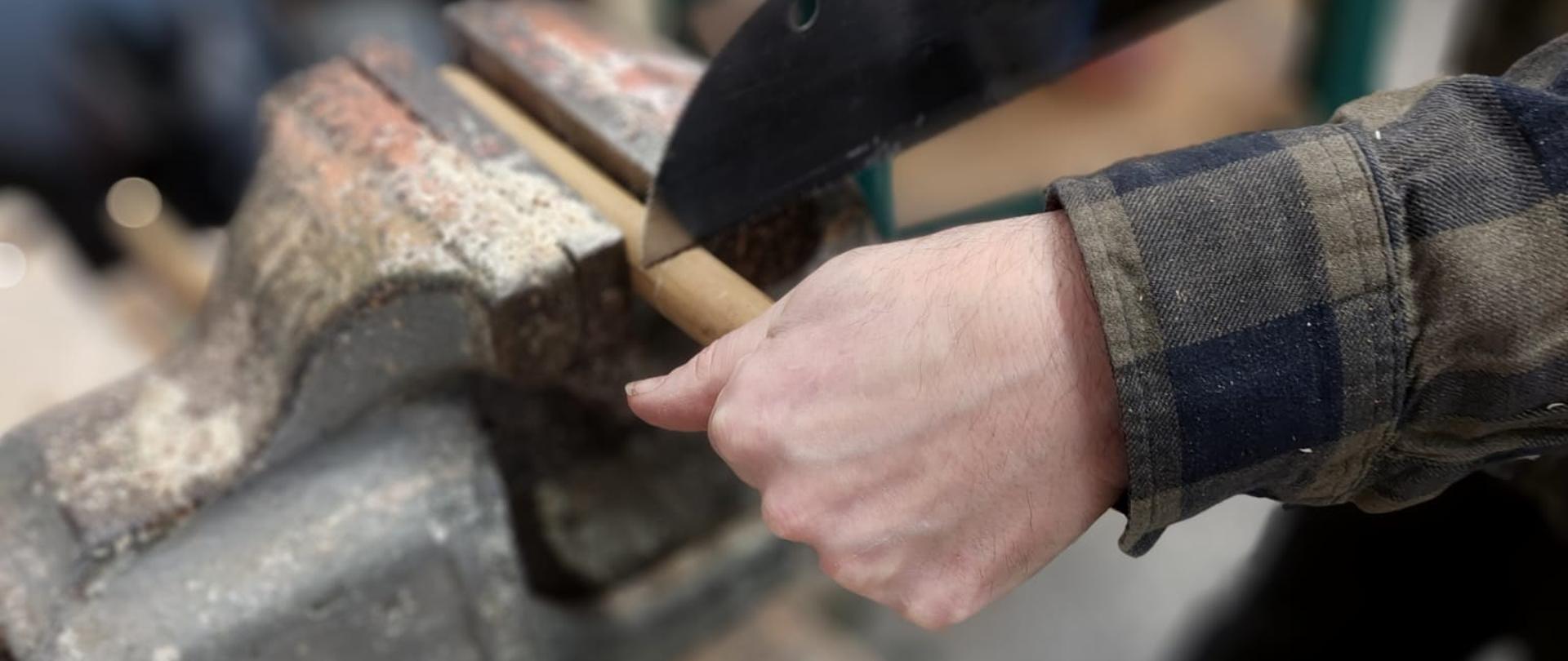 Osoba przecina drewniany patyk piłą ręczną, trzymając go w imadle na warsztatowym stole; wokół widać trociny i narzędzia.