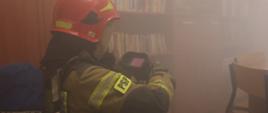 W pomieszczeniu wypełnionym dymem, przypominającym bibliotekę lub czytelnię, strażak w pełnym umundurowaniu ochronnym prowadzi działania z użyciem kamery termowizyjnej. Ubrany w kombinezon z oznaczeniem „PSP” oraz hełm i aparat oddechowy, funkcjonariusz przeczesuje wnętrze w poszukiwaniu źródeł zagrożenia lub osób wymagających ewakuacji. Ekran urządzenia ukazuje charakterystyczne różowe zabarwienie, wskazujące na obecność ciepła – kluczowy element w lokalizacji ognisk pożaru lub ludzi w trudnych warunkach widoczności.
Otoczenie, pełne regałów z książkami i rozstawionych krzeseł, podkreśla nietypowy charakter interwencji – działania prowadzone są w przestrzeni edukacyjnej, co wymaga szczególnej ostrożności i precyzji. Scena ukazuje nie tylko profesjonalizm służb ratowniczych, ale również znaczenie nowoczesnych narzędzi w skutecznym reagowaniu na zagrożenia.