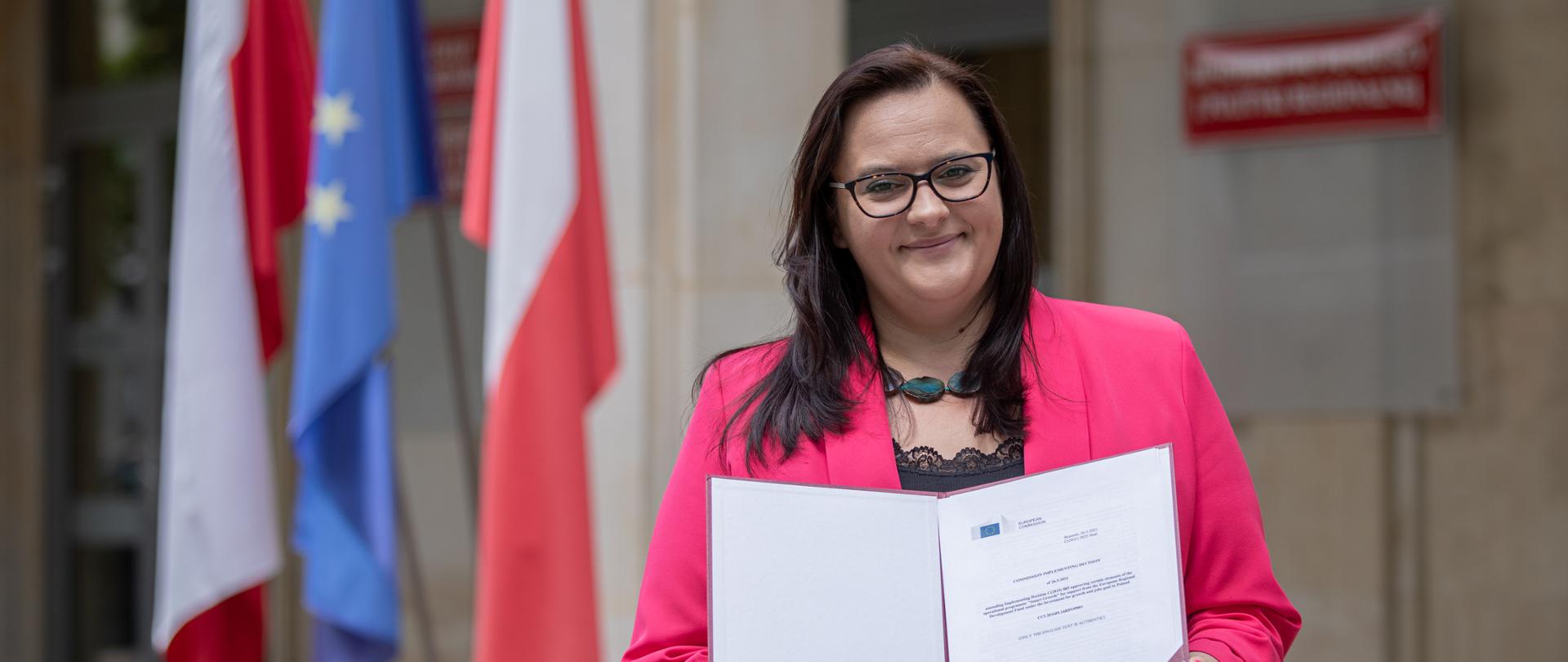 wiceminister Małgorzata Jarosińska-Jedynak trzyma przed sobą pozytywną decyzję Komisji Europejskiej za nią flagi Polski i Unii Europejskiej