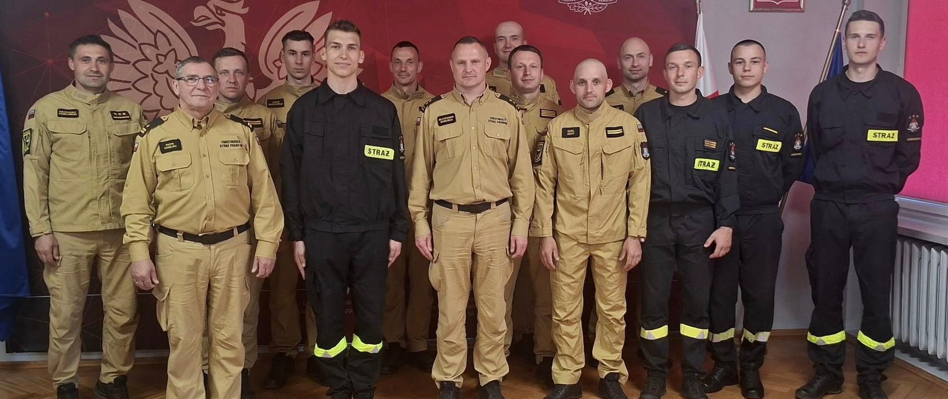 Oficjalne zdjęcie grupowe przedstawia czternastu funkcjonariuszy Państwowej Straży Pożarnej pozujących w pomieszczeniu służbowym na tle bordowej ścianki z białym orłem oraz napisem „Państwowa Straż Pożarna – Komenda Powiatowa PSP w Dzierżoniowie”. Mężczyźni stoją w kilku rzędach na jasnym parkiecie ułożonym w jodełkę, ubrani w regulaminowe umundurowanie: część z nich ma na sobie piaskowe mundury służbowe, a pozostali czarne ubrania koszarowe z charakterystycznymi żółtymi napisami „STRAŻ” i odblaskowymi pasami na nogawkach. W tle, po prawej stronie, widoczna jest polska flaga państwowa oraz zawieszone na ścianie godło w ramie, co podkreśla formalny i uroczysty charakter spotkania, związanego prawdopodobnie ze ślubowaniem strażackim.