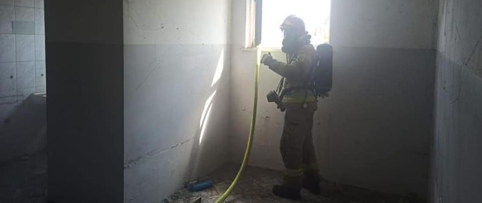 Widok na strażaka w aparacie ochrony dróg oddechowych. Strażak stoi przy oknie. Widok od wnętrza budynku.