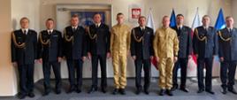 Grupa dziesięciu strażaków w sali ustawiona w rzędzie na tło z godłem Polski, gablotą oraz flagami państwowymi i służbowymi. Ośmiu strażaków nosi ciemnogranatowe mundury galowe ze złotymi sznurami, a dwóch w środku wyróżnia się piaskowymi mundurami. Wszystkie osoby stoją na baczność i patrzą w stronę aparatu.