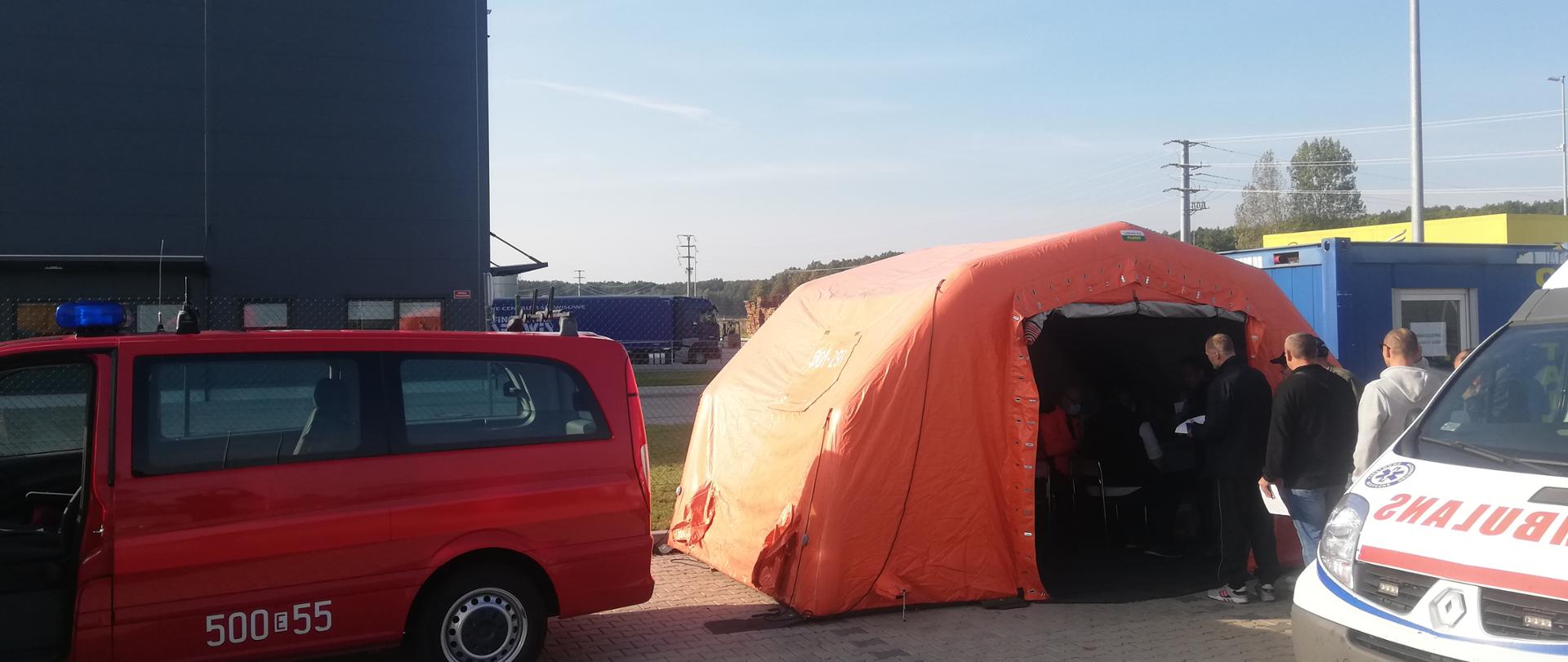 Po lewej stronie czerwony samochód straży pożarnej na tle zakładu K-FLEX Polska. Na środku zdjęcia znajduje się pomarańczowy namiot przed którym czekają osoby na szczepienie. Po prawej stronie znajduje się karetka pogotowia ratunkowego. 