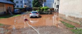 Zdjęcie przedstawia zalane podwórze przy budynkach mieszkalnych. W wodzie stoją dwa samochody osobowe. Na wodzie unosi się pompa pływająca z podłączonym wężem pożarniczym. 