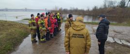 Strażak w mundurze musztardowym melduje mężczyźnie obok stoi zastępca komendanta głównego Państwowej Straży Pożarnej za nimi ustawieni są w dwóch rzędach strażacy za nimi płynie rzeka Wisła. 