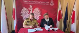 Oficjalne podpisanie umowy. Przy stole nakrytym czerwonym obrusem siedzą dwaj mężczyźni: po lewej strażak w piaskowym mundurze służbowym, po prawej mężczyzna z długą brodą w ciemnej marynarce. Obaj składają podpisy na dokumentach. W tle znajduje się czerwona ścianka z godłem Polski oraz napisem „PAŃSTWOWA STRAŻ POŻARNA”, a po bokach stoją flagi państwowe.