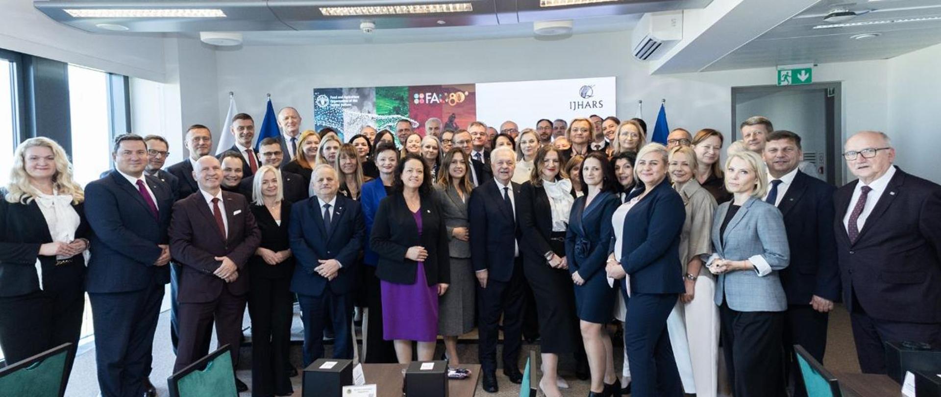 Zdjęcie grupowe kilkudziesięciu osób w eleganckich strojach biznesowych, stojących w kilku rzędach w jasnej sali konferencyjnej. Uczestnicy pozują do wspólnej fotografii, patrząc w stronę aparatu.