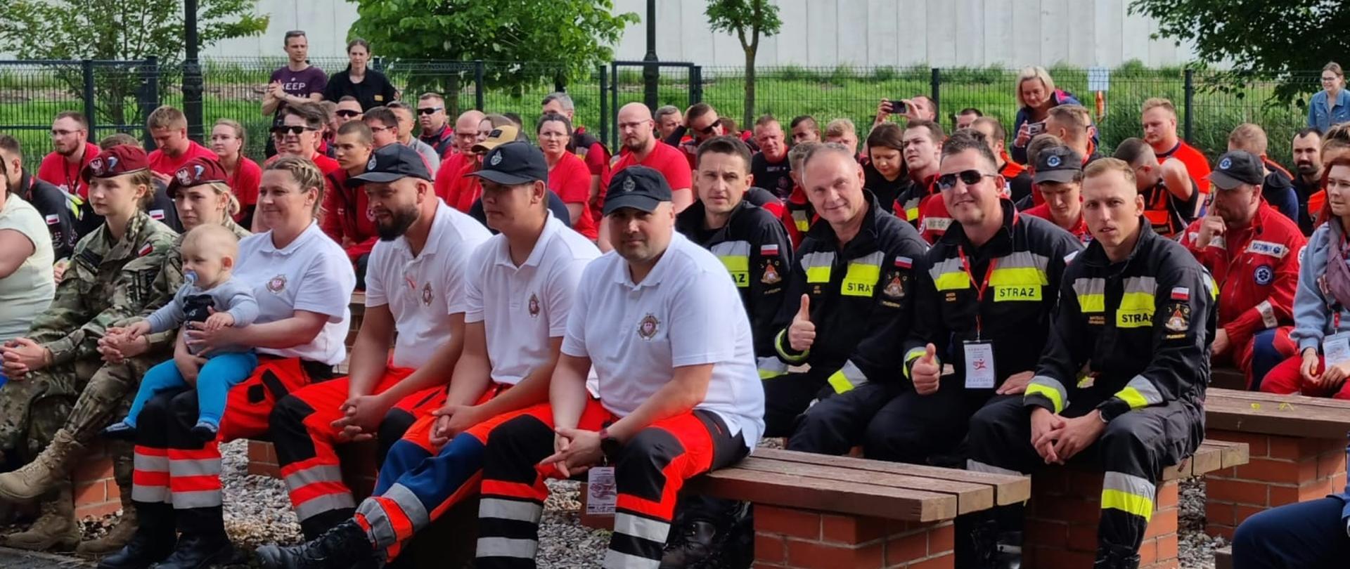 Kilkadziesiąt osób ubranych w mundury i stroje rtowników medycznych siedzi rzędami na drewnianych ławkach, wśród nich czterech zawodników ubranych w mundury PSP