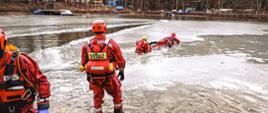 Ratownicy w specjalnych ubraniach ćwiczą na zamarzniętym akwenie. W tle obiekty i zabudowania kompleksu rekreacyjno-wypoczynkowego oraz las.