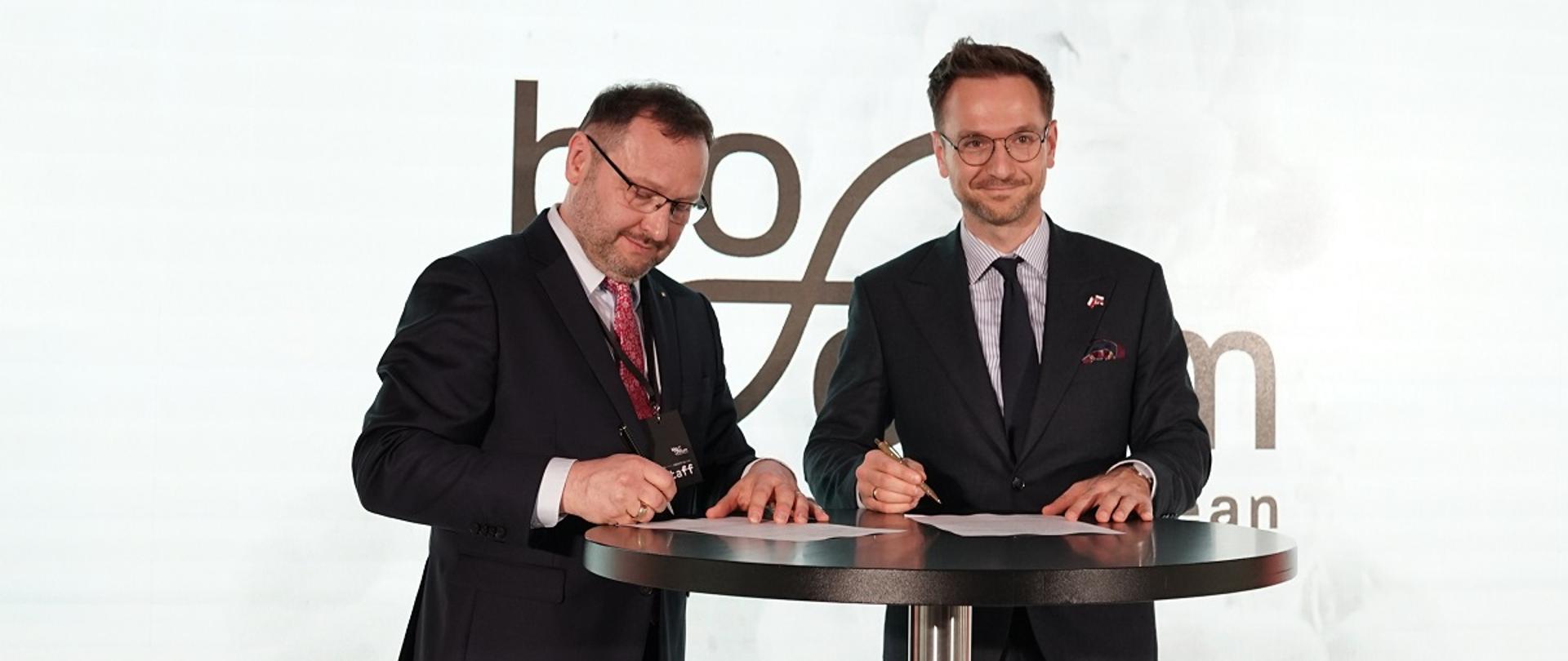 Minister rozwoju i technologii Waldemar Buda podczas podpisania deklaracji o współpracy między MRiT a Związkiem Związkiem Firm Biotechnolgicznych BioForum na rzez wsparcia sektora biotechnologii, minister Buda stoi przy stole i trzyma w ręku długopis, po jego lewej stronie deklarację podpisuje przedstawiciel ZFBB