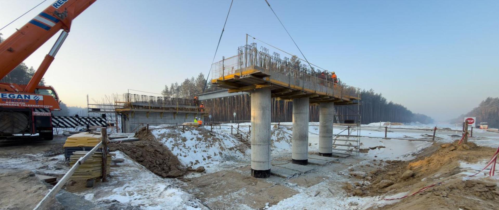 Plac budowy zimą - prace przy podporach obiektu mostowego. Montaż belki ustroju nośnego na podporach za pomocą dźwigu. W tle zaśnieżony ślad przyszłej drogi 