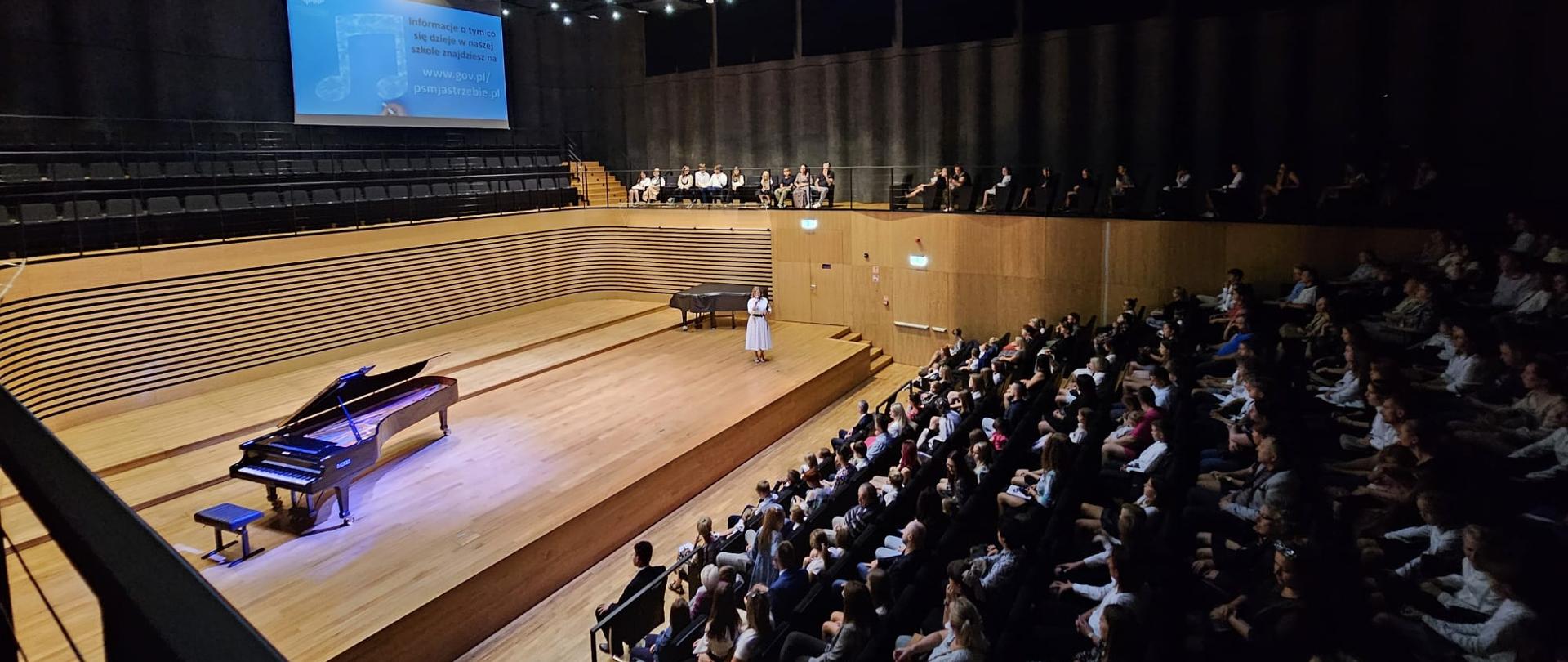 Widok z góry na widownię zapełniona publicznością i na estradę, na której stoi kobieta mówiąca do mikrofonu. Z boku estrady stoi fortepian. Nad sceną widoczny rzutnik z napisem "Informacje o tym co dzieje się w naszej szkole znajdziesz na www.gov.pl/psmjastrzebie.pl"