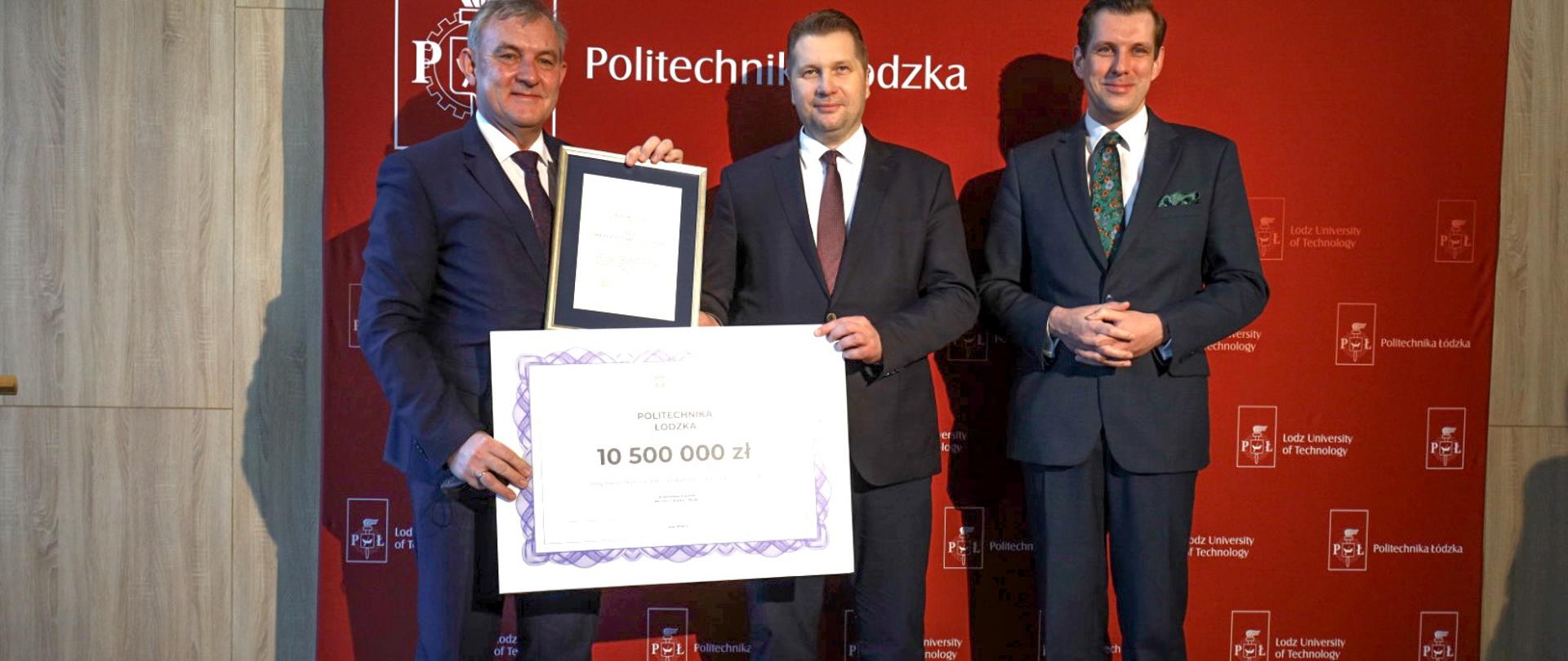 Minister Przemysław Czarnek pozuje do zdjęcia z czekiem oraz przedstawicielami Politechniki Łódzkiej, za nimi stoi baner z napisem Politechnika Łódzka.
