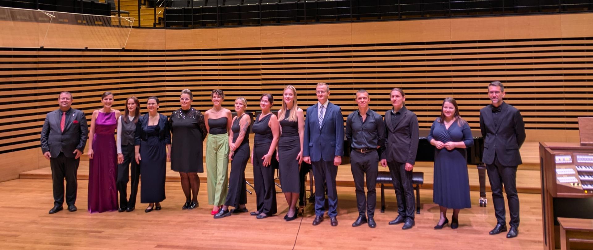 Czternaście osób stoi w rzędzie na estradzie sali koncertowej.