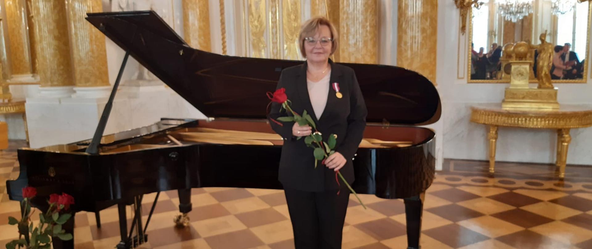 Zdjęcie przedstawia Panią Dorotę Chłopocką podczas uroczystości wręczenia złotego medalu za długoletnią służbę. W tle zdjęcia stoi fortepian.
