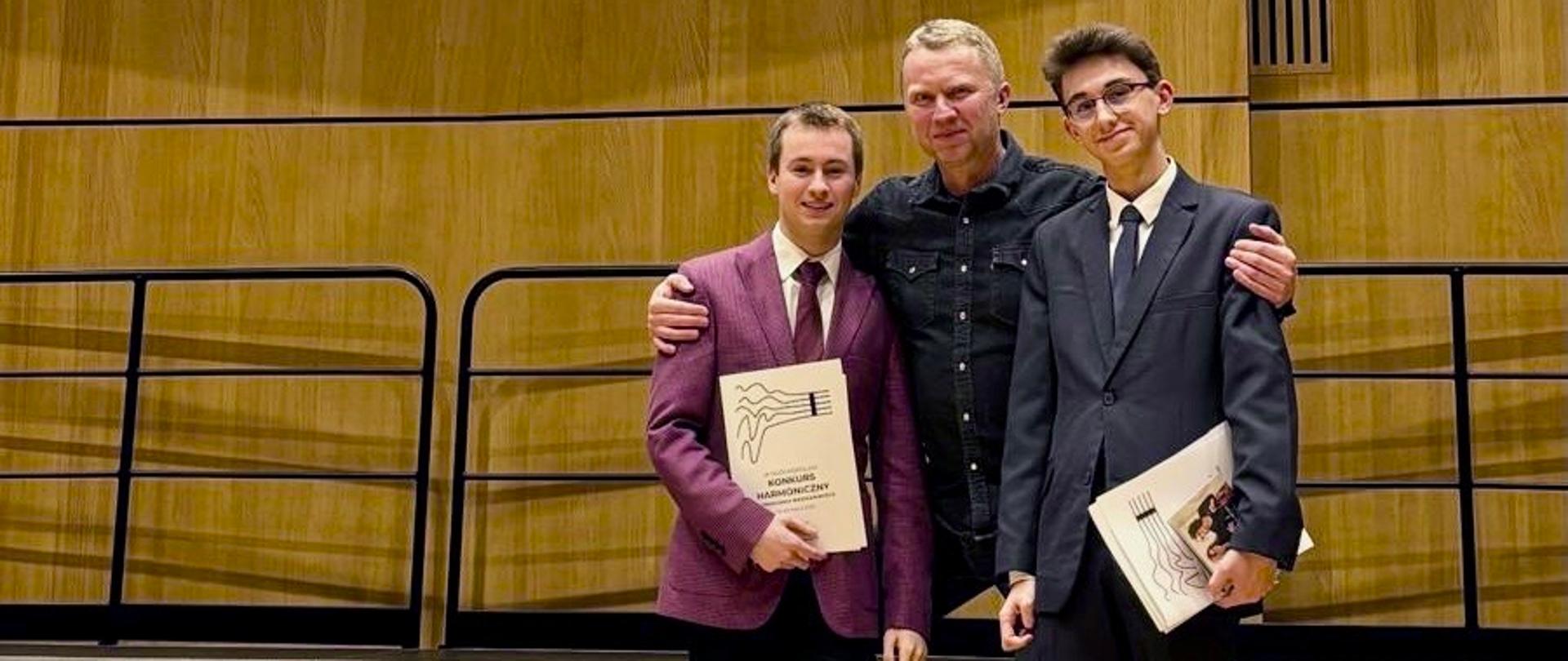 Kolorowe zdjęcie na schodach na scenie sali koncertowej w szkole muzycznej Łodzi Michała Galbierza, Gabriela Wojaczka i Pana Jacka Porszke. Wszyscy Panowie stoją obok siebie, w środku Pan Jacek, który trzyma uczniów za ramiona, natomiast uczniowie trzymają w rękach dyplom za zajęte miejsca.
