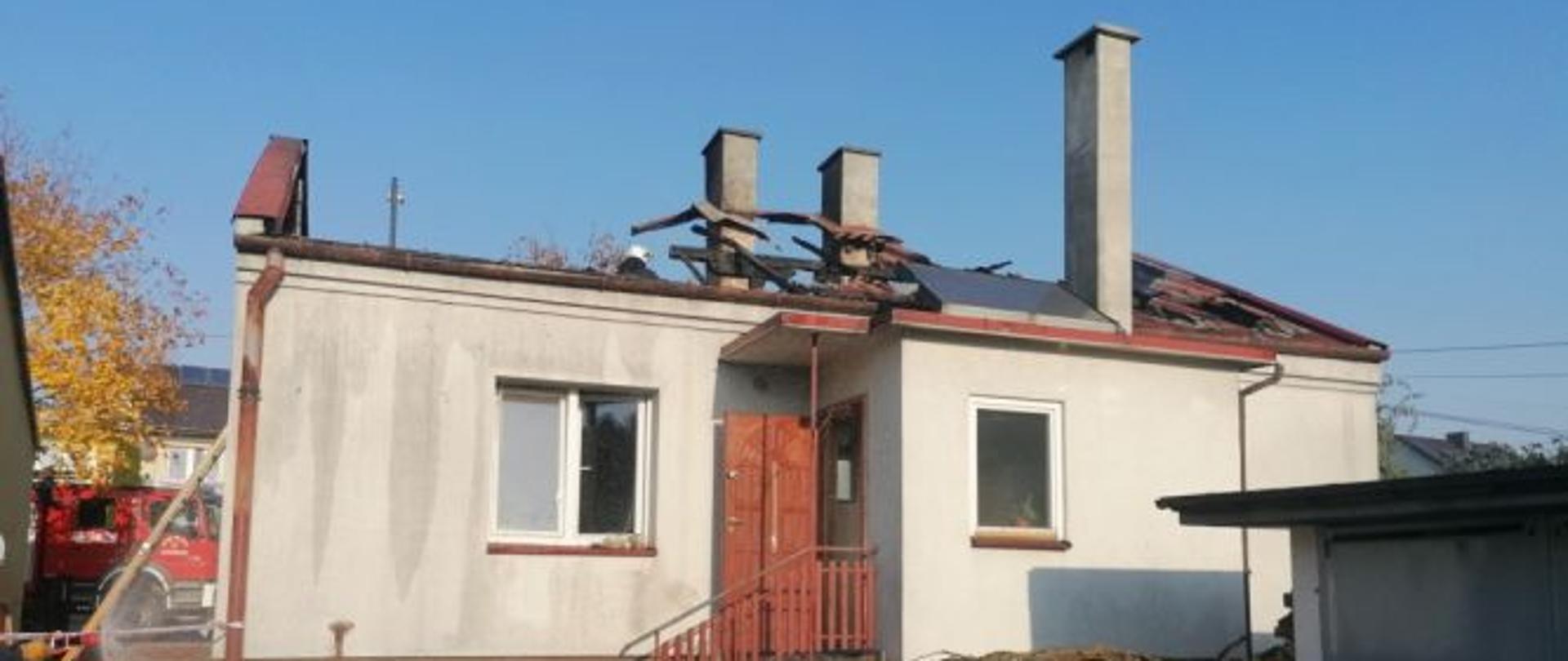 Na zdjęciu przedstawiony jest dom parterowy murowany z rozebraną więźbą dachową. Na trawniku ułożono nadpalone poszycie dachu. 