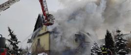 Widoczny pożar budynku mieszkalnego, silne zadymienie, podnośnik hydrauliczny i strażacy pracujący na nim