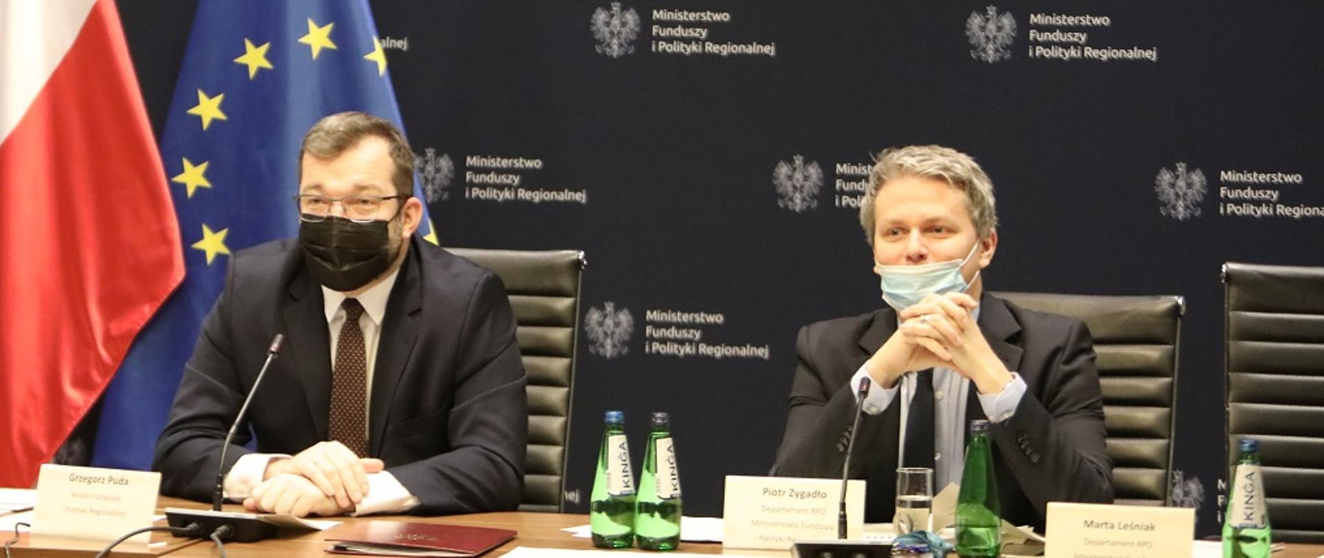Minister Grzegorz Puda i dyrektor Piotr Zygadło siedzą za stołem konferencyjnym. Za nimi ścianka z logo MFiPR oraz flagi Polski i UE.