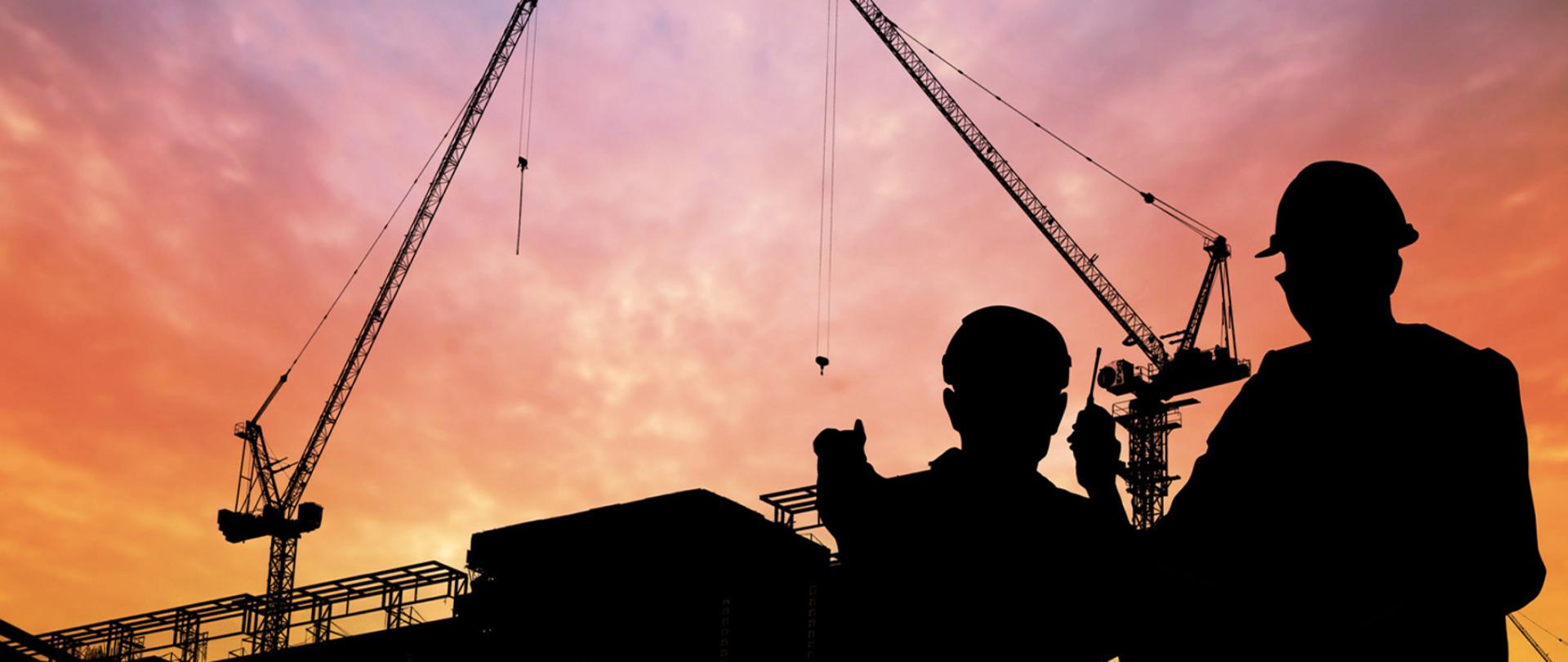 Dwóch pracowników w kaskach na tle placu budowy z żurawiami podczas zachodu słońca.