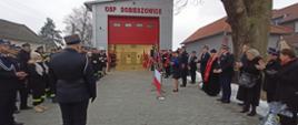 Widok od drogi w stronę remizy. W oddali w centrum remiza OSP Dobieszowice. Budynek w kolorze szarym i czerwoną bramą garażową podniesioną do połowy. Po prawej trybuna honorowa oraz zaproszeni goście. Po lewej pododdział strażaków w mundurach wyjściowych i koszarowych. Po lewej widać też od tyłu stojącego dowódcę uroczystości. 