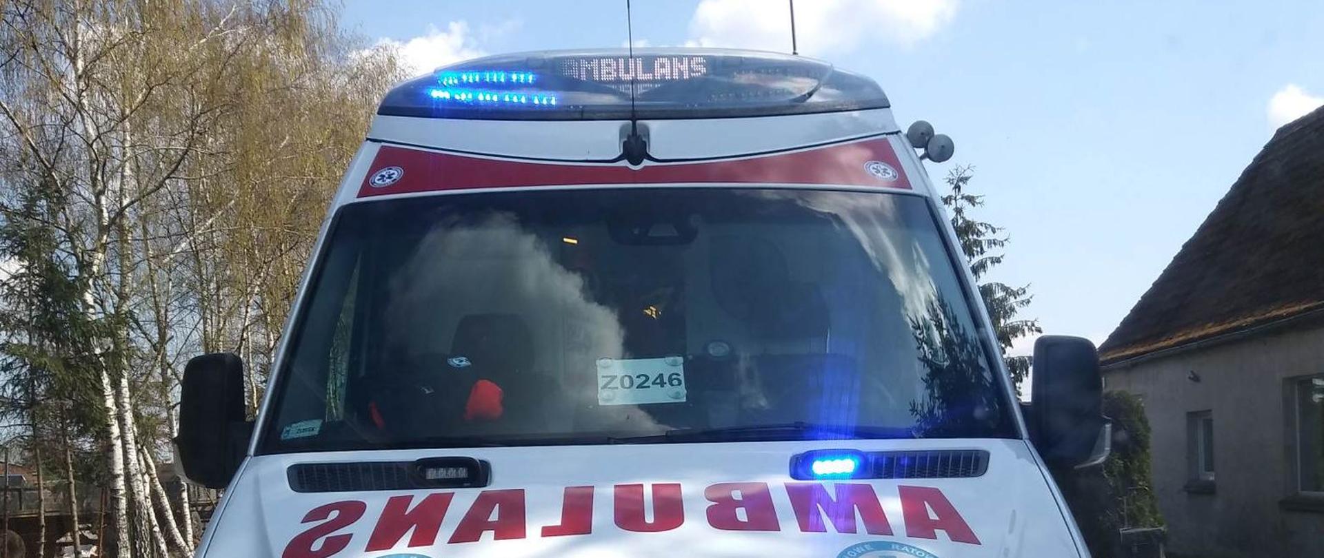 Zdjęcie przedstawia ambulans w tle widzimy drzewa oraz stare budynki.