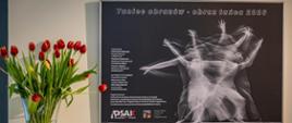 Zdjęcie przedstawia plakat do wystawy taniec obrazów obraz tańca. Po lewej stronie stoi wazon z czerwonymi tulipanami.