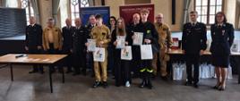 Trójka starszych uczestników stoi z przodu, trzymając dyplomy i torby z nagrodami, pozując do zdjęcia w dużej, eleganckiej sali z wysokimi oknami i zasłonami. Jedna osoba ubrana jest w strój strażacki. Za nimi ustawiona jest grupa dorosłych, w tym osoby w mundurach strażackich. W tle widoczne są banery Ochotniczej i Państwowej Straży Pożarnej, co wskazuje na uroczyste wręczenie nagród najstarszej grupie uczestników konkursu o tematyce pożarniczej.