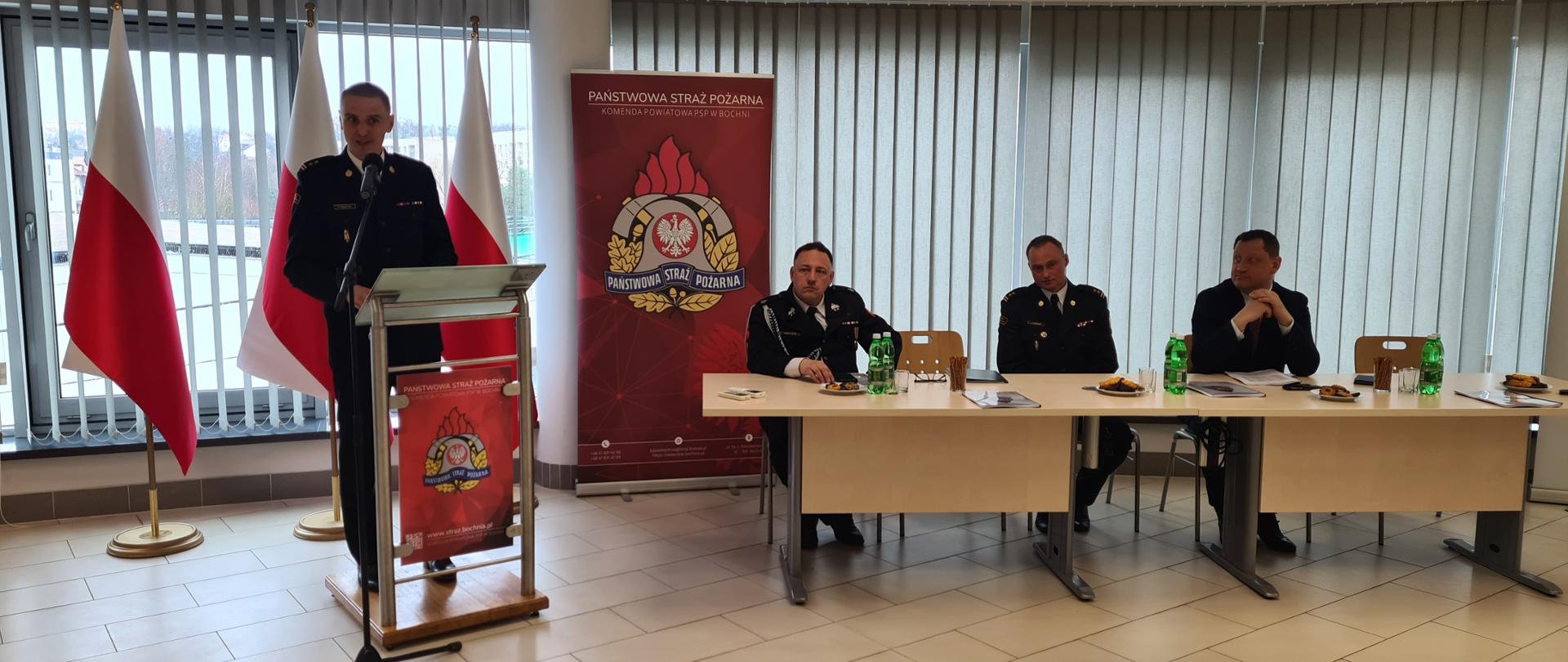 Zdjęcie ukazuje w centralnej części strażaka – Zastępcę Małopolskiego Komendanta Wojewódzkiego PSP ubranego w strój strażacki wyjściowy, który stojąc na mównicy wygłasza przemówienie. Na drugim planie siedzą dwóch strażaków oraz jeden mężczyzna ubrany w garnitur. W tle znajduje się rolety koloru szarego oraz flagi państwowej koloru biało-czerwonego
