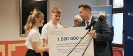 Minister Czarnek stoi przed dwoma mikrofonami na stojakach trzyma wielki symboliczny czek z napisem 1 500 000 zł, obok niego uczeń i uczennica. W tle na ścianie obraz.