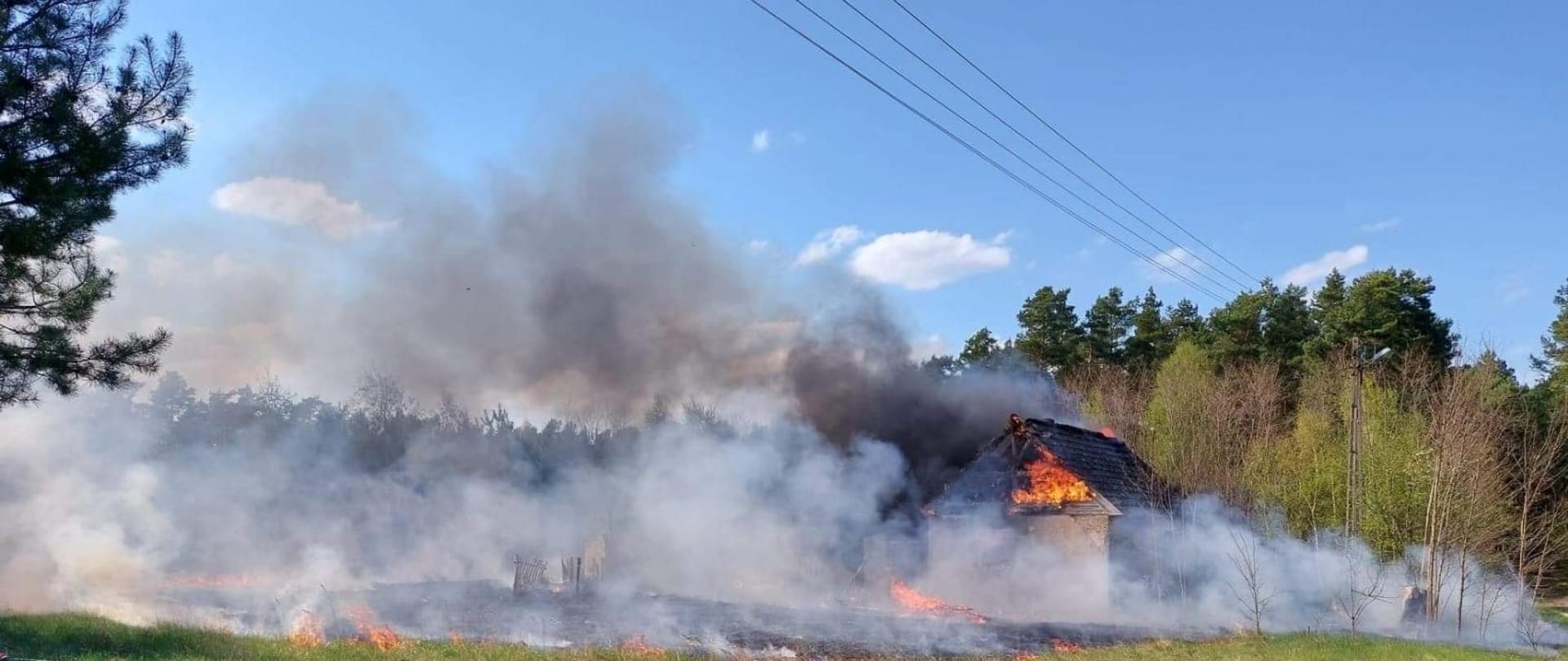 Na pierwszym planie skrzynka elektryczna. W tle płonący pustostan i okoliczne nieużytki. Za budynkiem pobliski las.