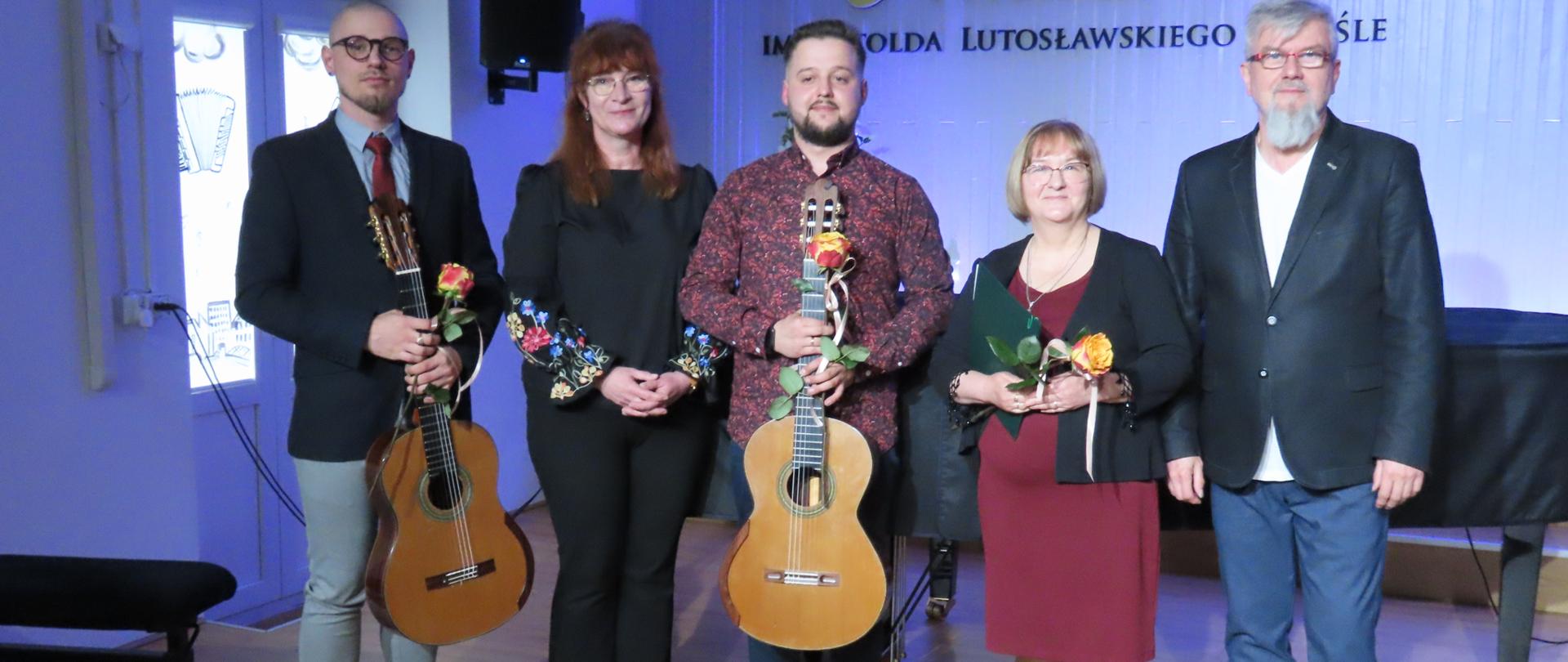 Na zdjęciu trzech mężczyzn pomiędzy którymi stoją dwie kobiety. Wszyscy w różnokolorowych ubraniach. Dwaj mężczyźni trzymają w rękach gitary i kwiaty, niższa z kobiet też trzyma w rękach kwiat. Za nimi jasna ściana, na której wisi godło państwowe i logo szkoły z napisem Państwowa Szkoła Muzyczna I stopnia im. Witolda Lutosławskiego w Jaśle. Z prawej i lewej strony stojących stoją ławy fortepianowe, a z przodu widać dwa podnóżki gitarowe.
