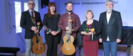 Na zdjęciu trzech mężczyzn pomiędzy którymi stoją dwie kobiety. Wszyscy w różnokolorowych ubraniach. Dwaj mężczyźni trzymają w rękach gitary i kwiaty, niższa z kobiet też trzyma w rękach kwiat. Za nimi jasna ściana, na której wisi godło państwowe i logo szkoły z napisem Państwowa Szkoła Muzyczna I stopnia im. Witolda Lutosławskiego w Jaśle.