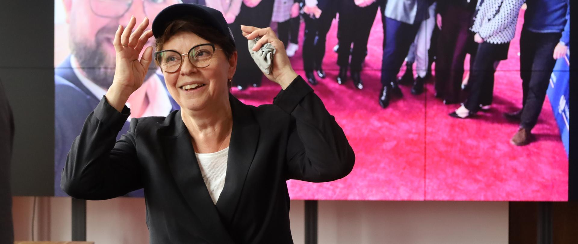 Kobieta stoi przed ekranem. Na głowie ma czapkę. Za nią zdjęcie współpracowników.