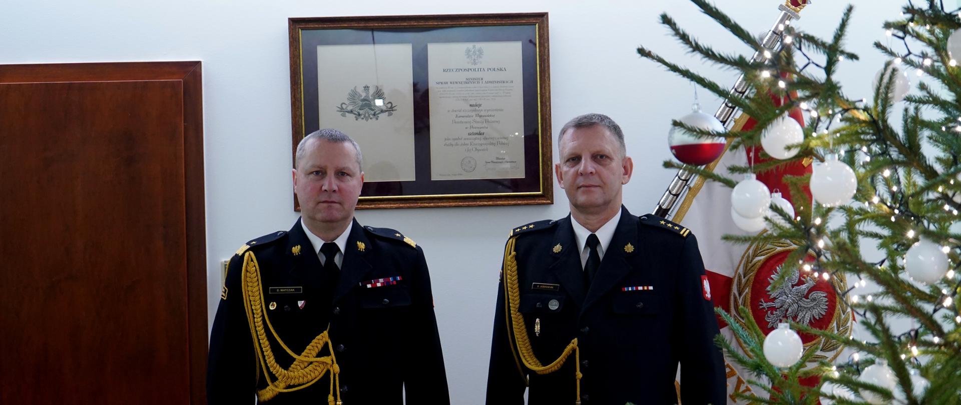 dwóch strażaków w mundurach ze sznurem stoi obok siebie, w tle sztandar i choinka 