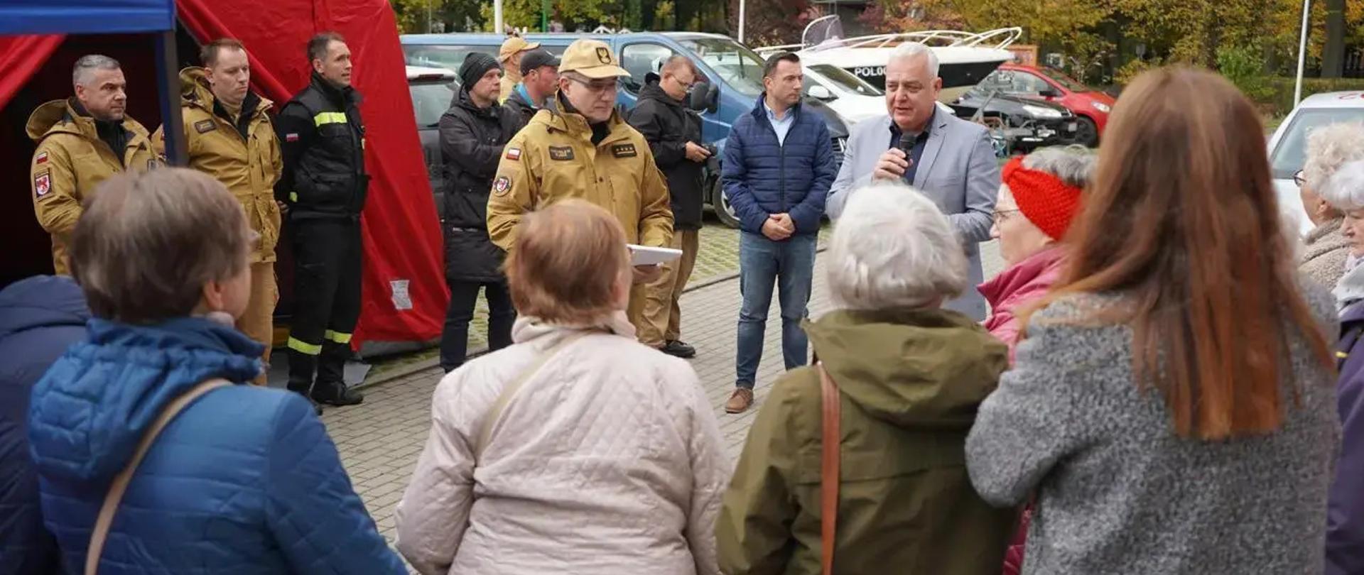 Na zdjęciu widoczna jest scena z wydarzenia plenerowego akcji „Czujne Zachodniopomorskie”, w którym uczestniczą funkcjonariusze straży pożarnej oraz mieszkańcy, głównie osoby starsze. Na pierwszym planie widać grupę seniorów uważnie słuchających przemówienia mężczyzny wice starosty szczecineckiego Pana Daniela Raka w jasnoszarej marynarce, który trzyma mikrofon i przemawia do zebranych. Obok niego stoi strażak komendant powiatowy starszy kapitan Mirosław Śledź w mundurze koloru piaskowego, trzymający notatki, a za nim kilku innych funkcjonariuszy w ubraniach strażackich – zarówno w kolorze czarnym, jak i piaskowym.
W tle widać czerwony namiot oraz zaparkowane pojazdy, w tym policyjną łódź i samochody służbowe. Wydarzenie odbywa się na brukowanym placu otoczonym drzewami, które mają jesienne barwy – żółcie, czerwienie i zielenie – co sugeruje, że spotkanie ma miejsce jesienią. Atmosfera jest spokojna i edukacyjna, co wskazuje na charakter informacyjny lub profilaktyczny wydarzenia, prawdopodobnie dotyczącego bezpieczeństwa przeciwpożarowego.
