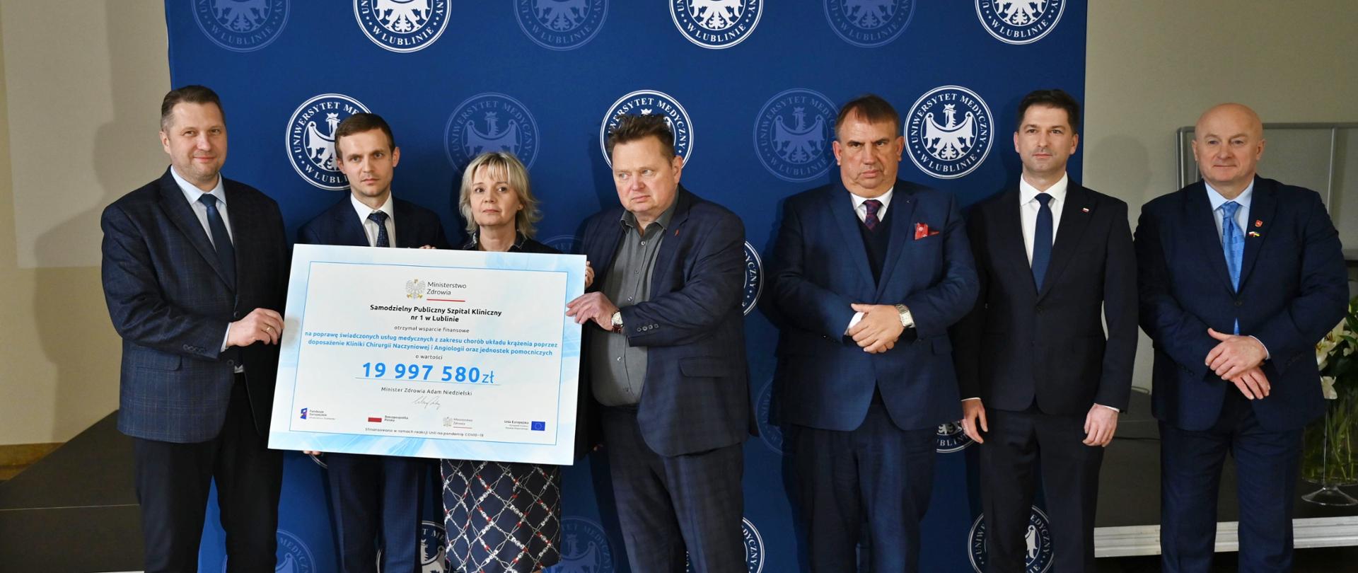 Na podłodze z szarożółtych kamiennych kafli stoi siedem elegancko ubranych osób - sześciu mężczyzn w garniturach i kobieta w spódnicy w granatowo-szarą kratę, dwóch trzyma wielki symboliczny czek z napisem 19 997 580 zł, za nimi granatowa ścianka stojąca na tle żółtej ściany.