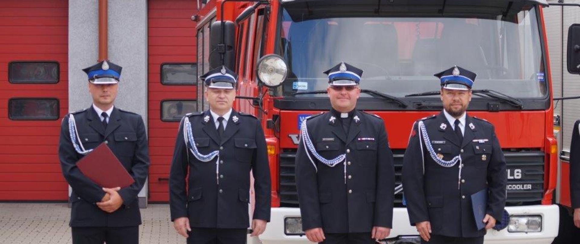 Czterech strażaków ubranych w mundury wyjściowe stoi w szeregu na baczność na tle samochodu ratowniczo-gaśniczego. Dwóch z nich w dłoniach trzyma teczki z dokumentami.