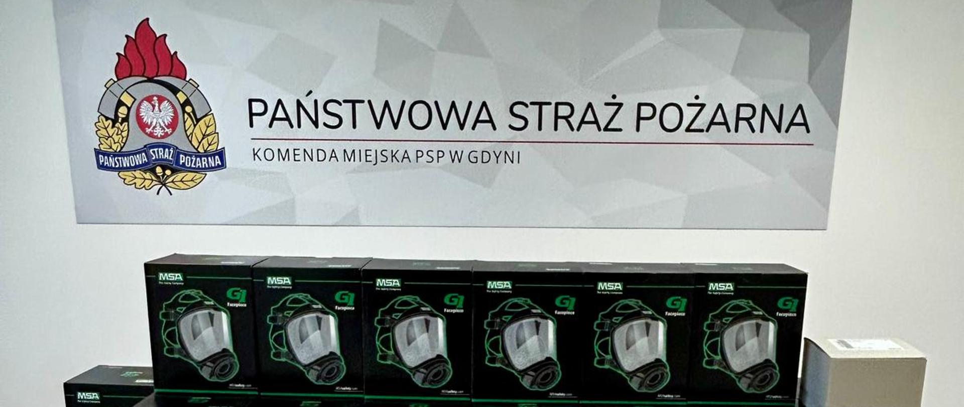 Maski i łączność radiowa ustawiona w pudełkach na podłodze. W tle na ściana napis i logo Państwowej Straży Pożarnej