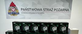 Maski i łączność radiowa ustawiona w pudełkach na podłodze. W tle na ściana napis i logo Państwowej Straży Pożarnej