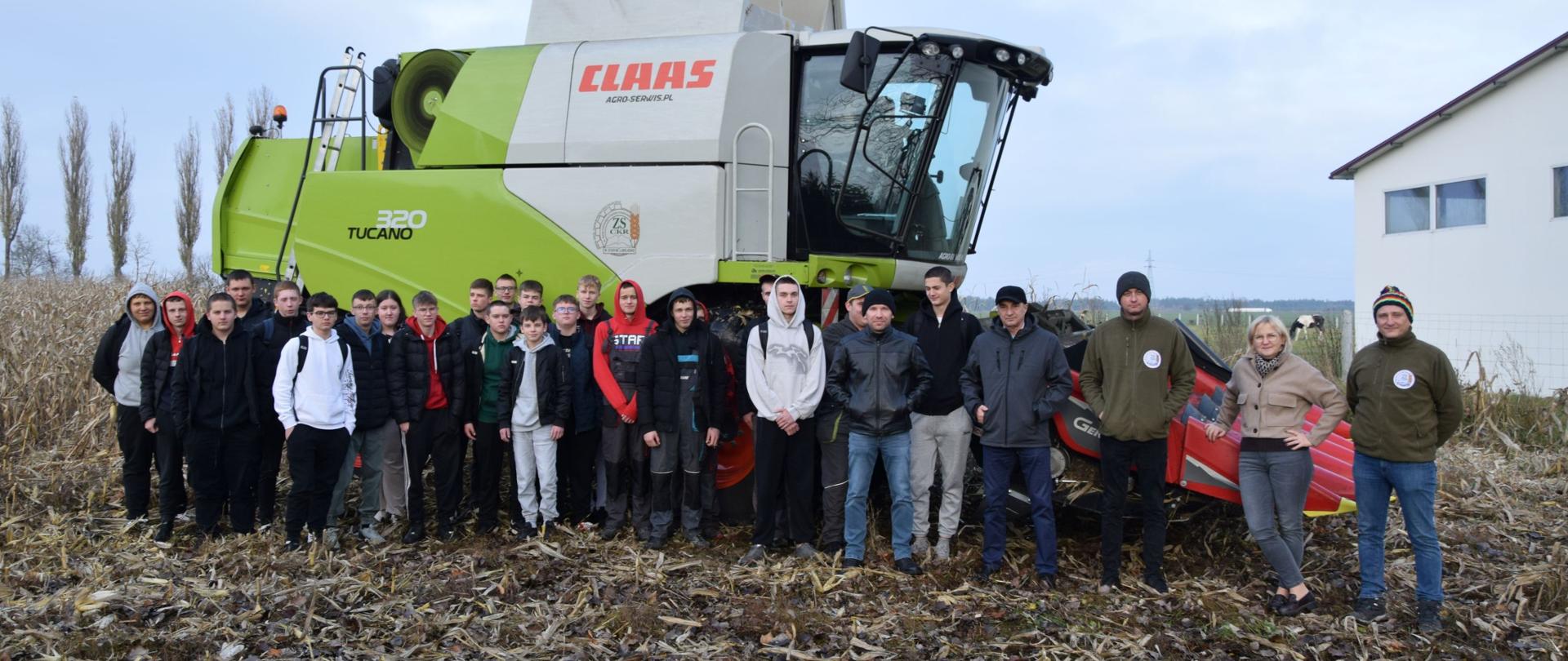 Grupa młodzieży biorąca udział w zbiorze wraz z opiekunami i kierownictwem szkoły na polu po zebranej kukurydzy, za nimi kombajn z logo szkoły