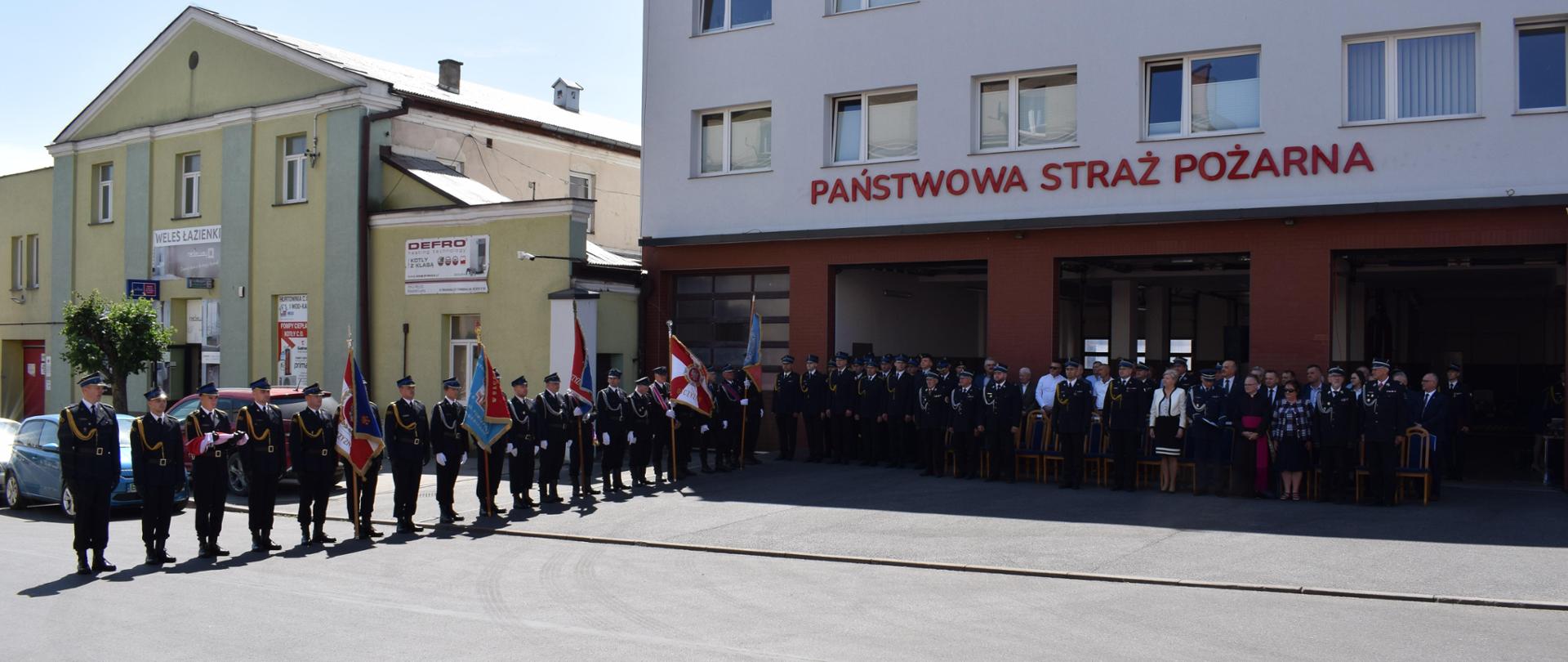 przed budynkiem straży na baczność stoją strażacy ubrani w mundury i ksiądz i kilka osób cywilnych z boku w szeregu stoją strażacy zze sztandarami