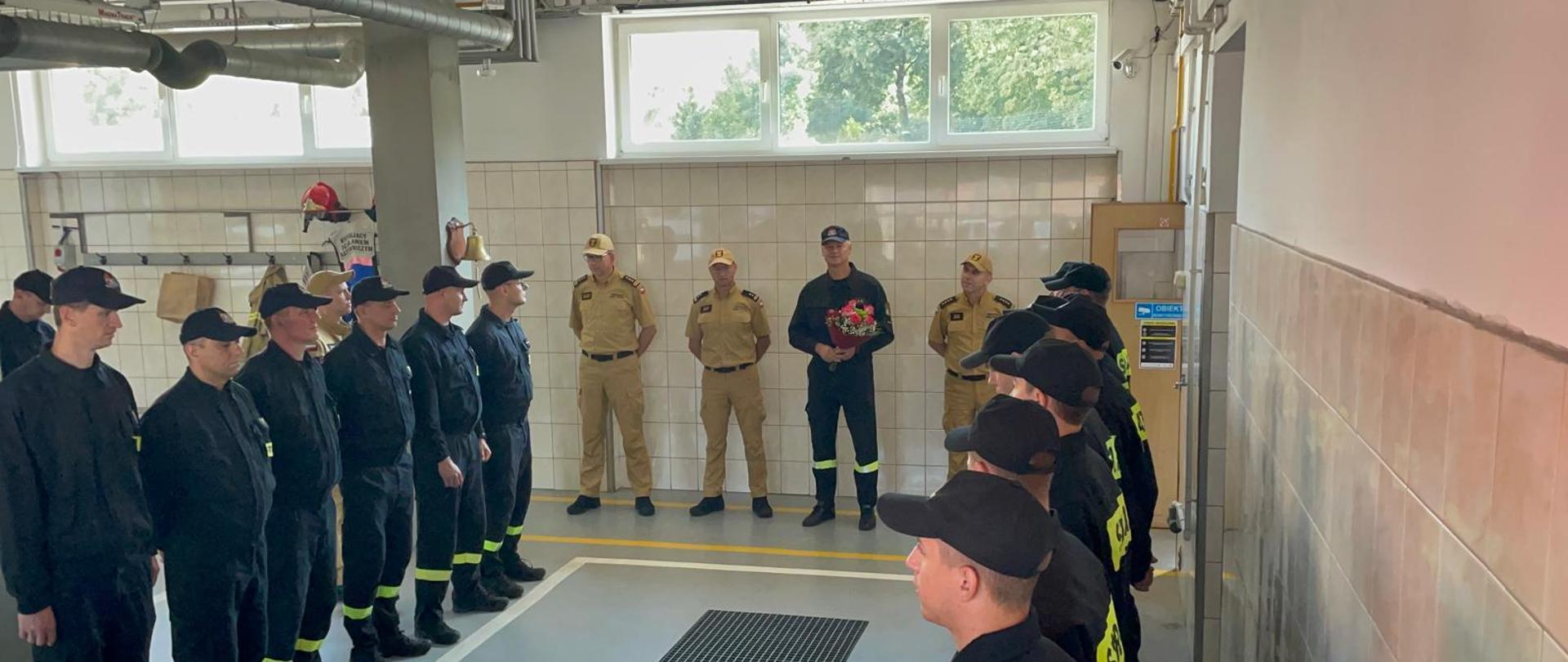 W oświetlonym garażu widoczne dwa rzędy stojących na przeciw siebie strażaków, z tyłu widoczni stojący obok siebie strażacy jeden z nich trzyma w ręku czerwone kwiaty, 