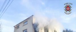 Zdjęcie przedstawia dwupiętrowy budynek i dym wydobywający się z dwóch okien na pierwszym piętrze