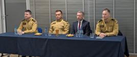 Czterech mężczyzn siedzi przy stole konferencyjnym podczas rocznej odprawy służbowej PSP w Myszkowie. Trzech z nich ma na sobie umundurowanie strażackie, jeden garnitur. Na stole stoją butelki z wodą, szklanki oraz drobny poczęstunek. W tle widoczne są rolety okienne i jasne ściany sali konferencyjnej.