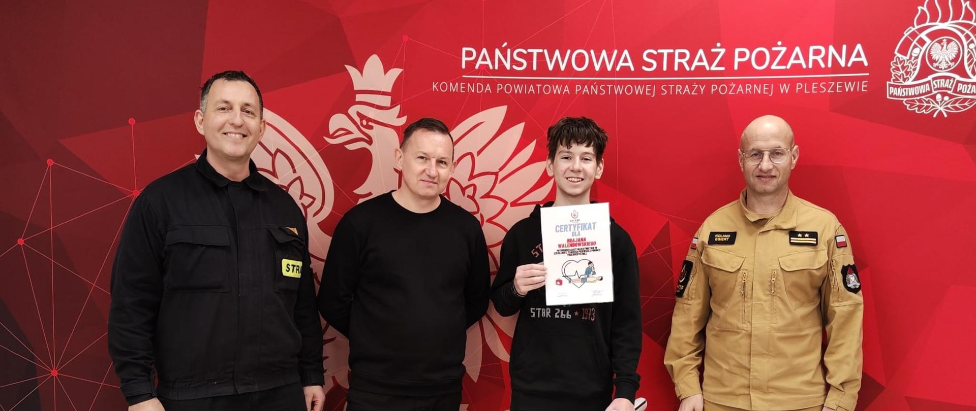 Na tle banera państwowej straży pożarnej stoją strażacy oraz mężczyzna i chłopak z certyfikatem.